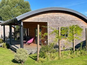 A wooden cabin with a curved roof, a covered patio with chairs, and a small garden area.