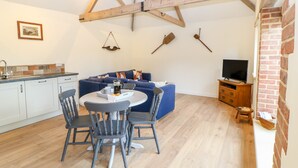 A modern kitchen with white cabinets, a dining table with chairs, a blue sofa, and a television on a wooden stand. - The Granary (Dereham)