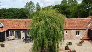A courtyard with a weeping willow tree, a wooden bench, and a small garden area with potted plants. - The Stables (Dereham)