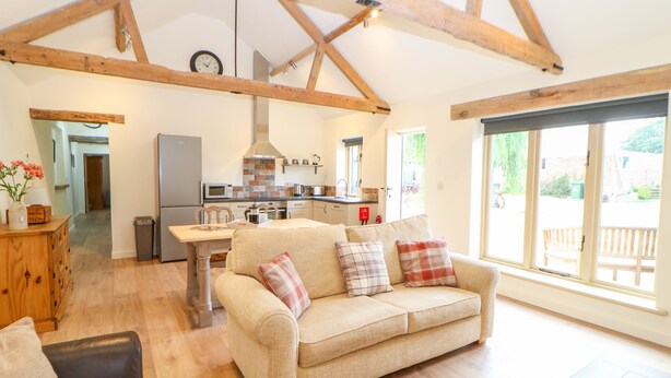 A spacious living room with a high ceiling and exposed wooden beams, a kitchen with modern appliances, and large windows offering a view of the outdoors. - The Stables (Dereham)