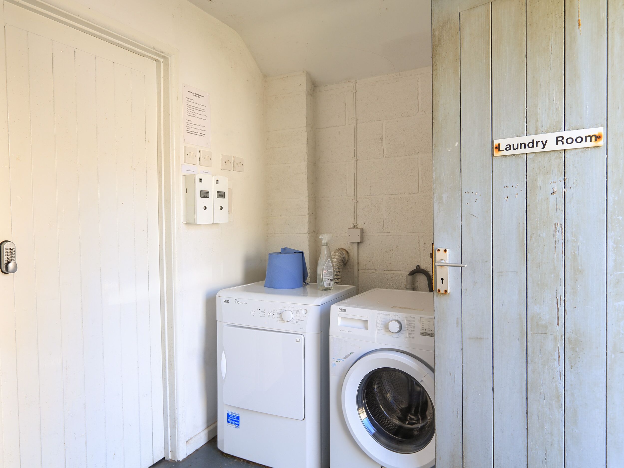 A laundry room with two washing machines, a door with a sign, and electrical outlets on the wall.