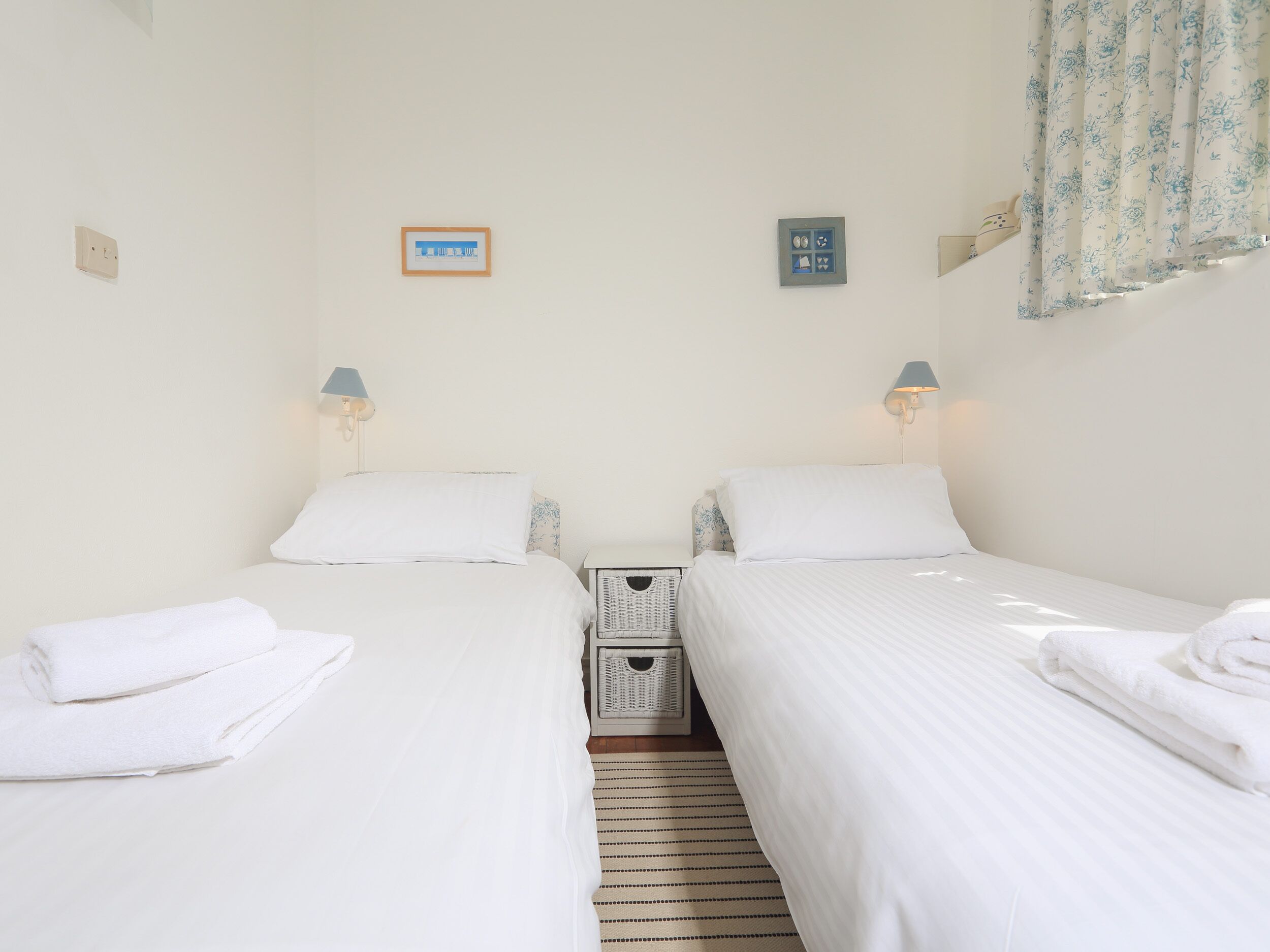 A twin room with two beds, each with a pillow and a lamp. There is a small basket on the floor and a wall-mounted light switch.
