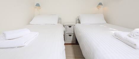 A twin room with two beds, each with a pillow and a lamp. There is a small basket on the floor and a wall-mounted light switch.