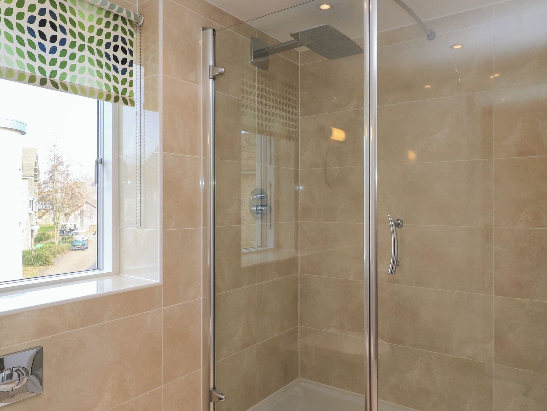 A glass-enclosed shower with beige tiles and a window with a green patterned blind.
