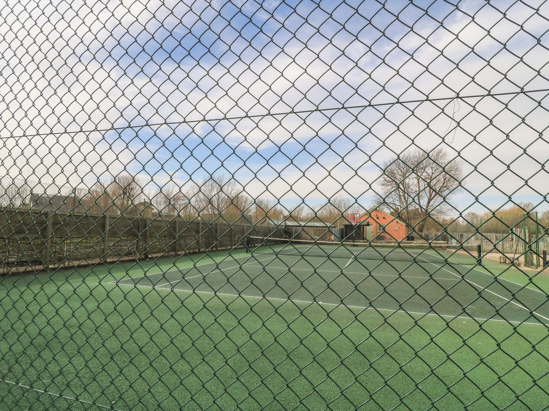A tennis court enclosed by a chain-link fence with a basketball hoop in the background.