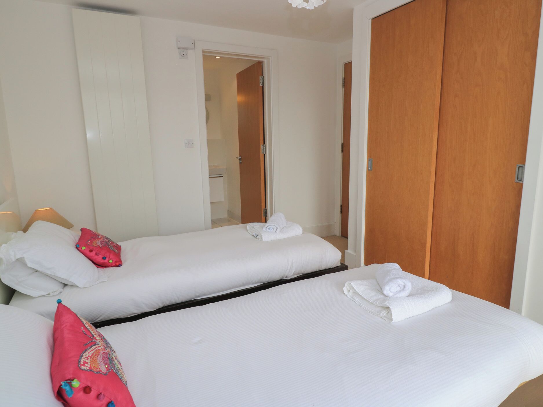 A hotel room with two beds, each with white linens and a pink decorative pillow. There is a wooden wardrobe and a mirror on the wall.