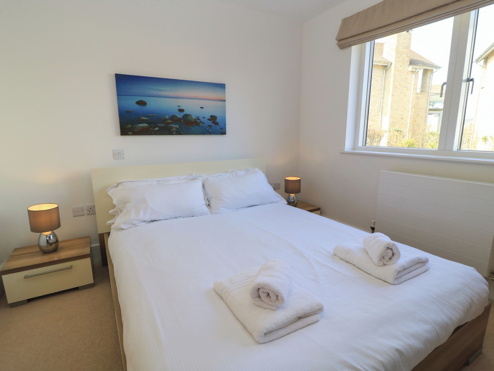 A neatly made bed with white linens and two folded towels, a painting of a landscape on the wall, and a window with a view of buildings outside.