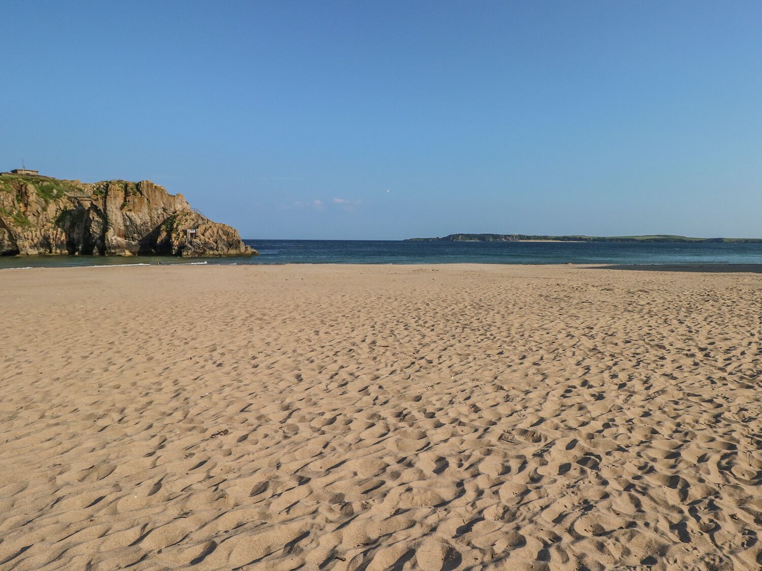 Una amplia playa de arena con huellas, un acantilado rocoso y un mar tranquilo bajo un cielo despejado.