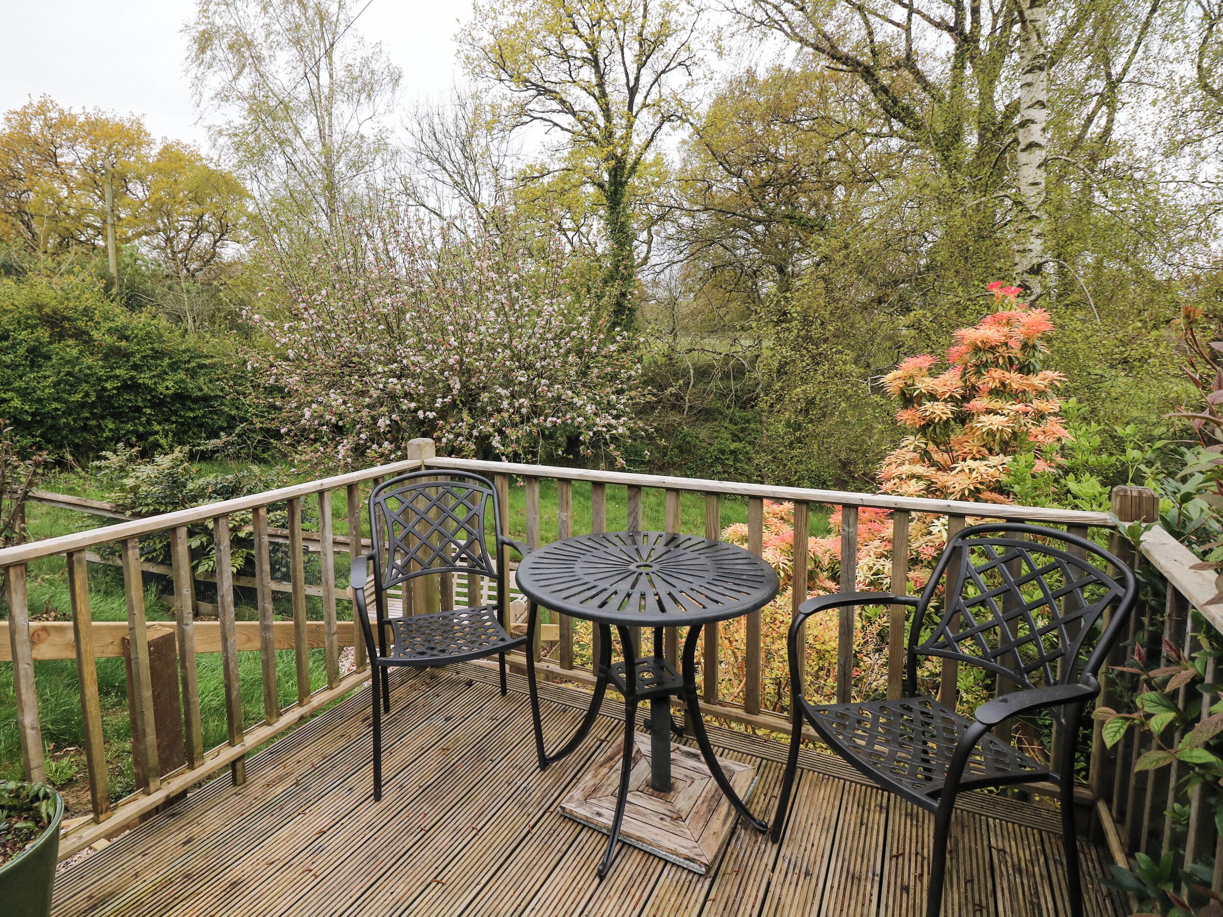 A wooden deck with a table and chairs, surrounded by a wooden fence and various trees and shrubs.