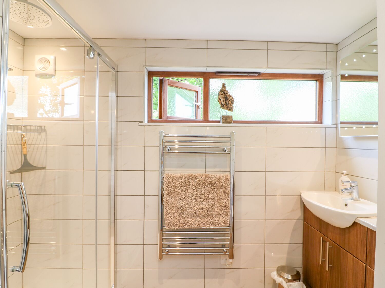 Un baño moderno con cabina de ducha de vidrio, un perchero con una toalla color beige, una ventana con vistas a la vegetación y un tocador de madera con un lavabo.