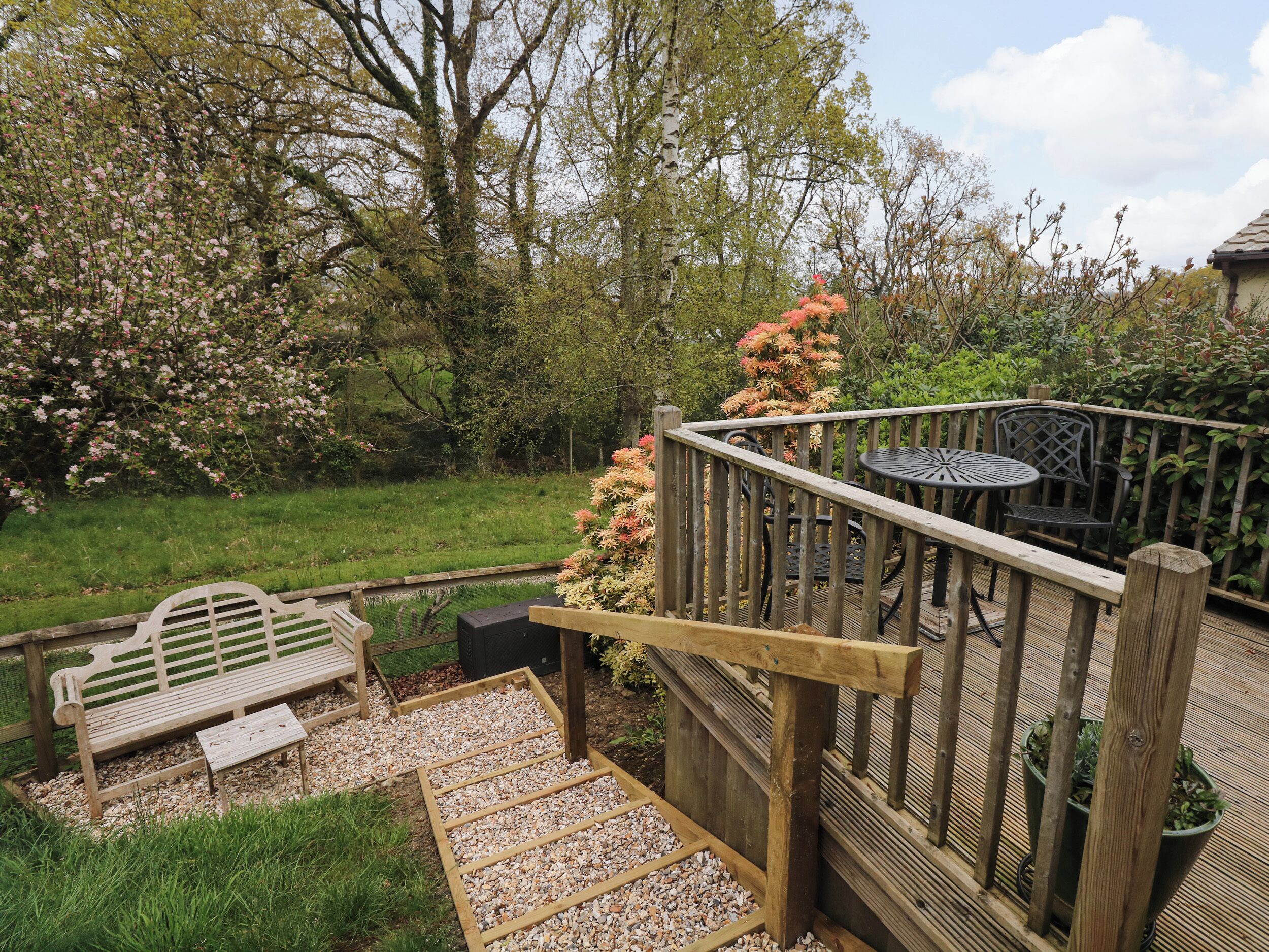 A wooden deck with a table and chairs, surrounded by a garden with trees and a bench.