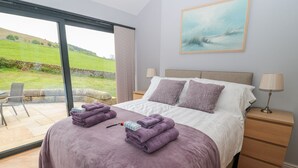 A bedroom with a large bed, two bedside tables, a nightstand, a chair, and a view of a green landscape through a glass door. - Coal Drops (Alnwick)