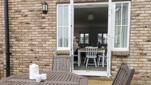 An outdoor patio with a wooden table and chairs, a brick wall, and a door leading to an interior dining area. - Causeway Cottage (Filey)