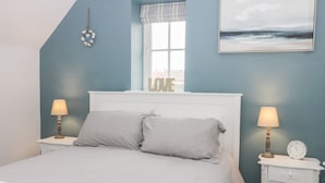 A bedroom with a bed, two bedside tables with lamps, a window with a view, and a wall-mounted painting. - Causeway Cottage (Filey)