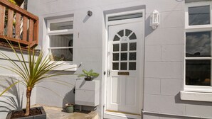 Un edificio blanco con una puerta marcada '12', un ventanal pequeño y una planta en maceta al frente.