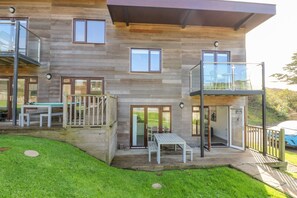 Una casa moderna de dos pisos con terraza, asientos exteriores y barandilla de vidrio.