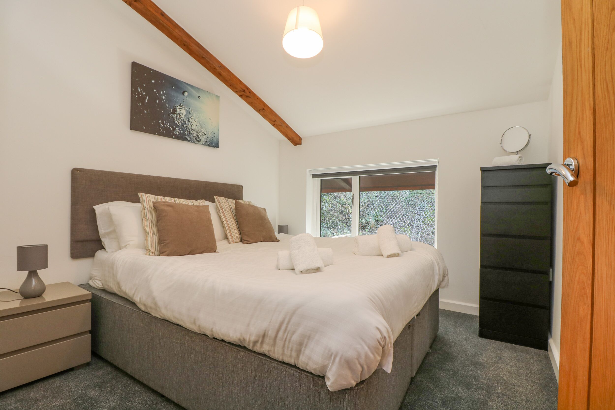 A bedroom with a large bed, a nightstand, a bedside lamp, a window with a view, and a door.
