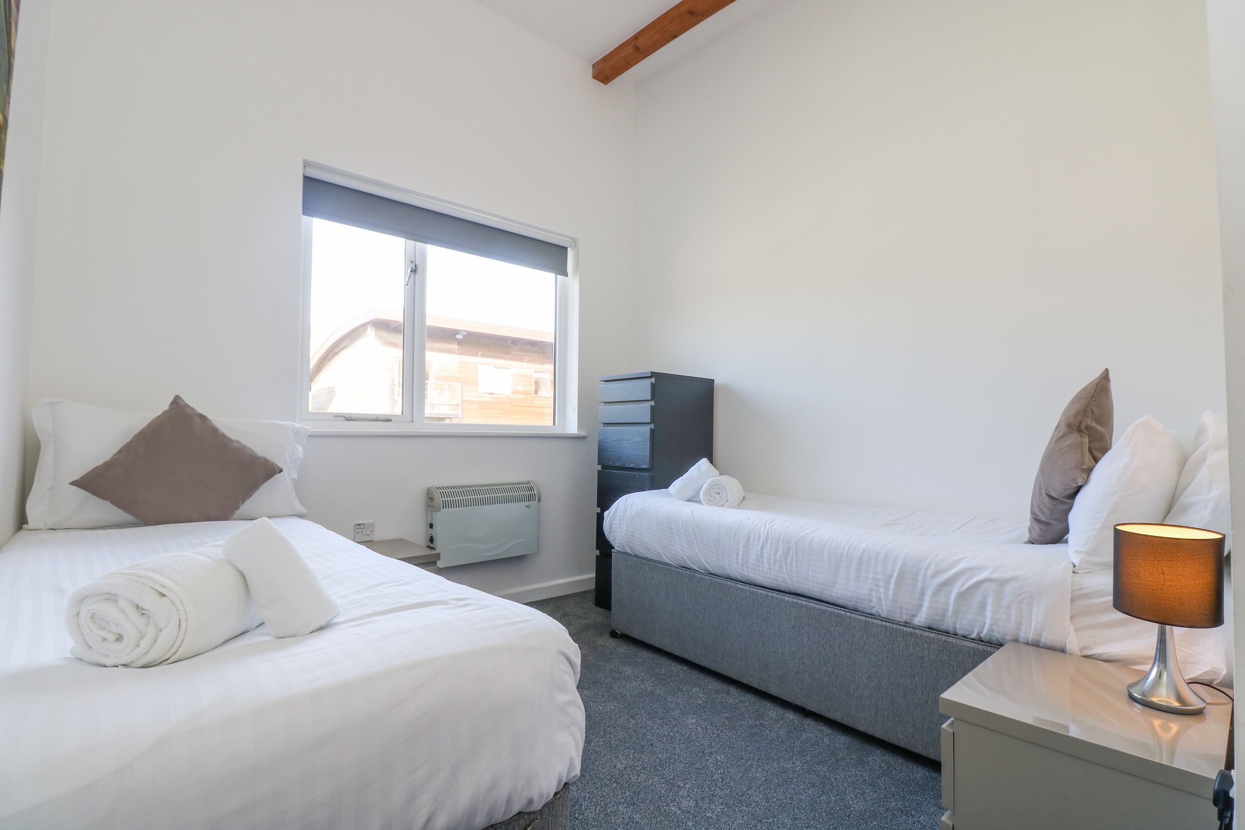 A bedroom with two beds, a window, a heater, and a bedside table with a lamp.