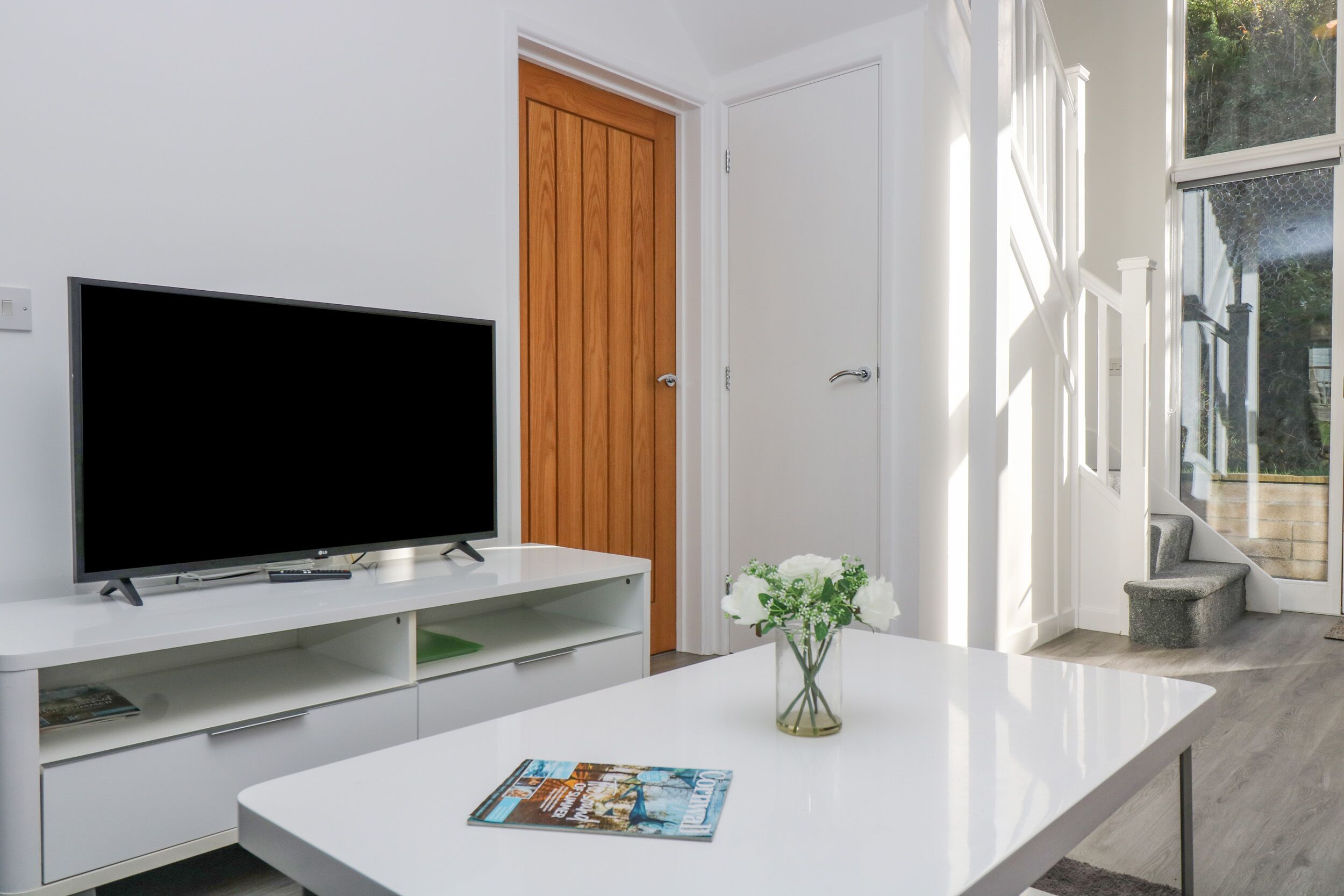 A modern living room with a flat-screen TV on a white console, a wooden door, and a glass door leading to a balcony.