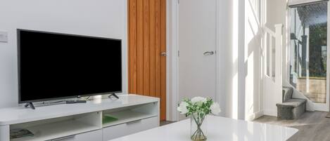 A modern living room with a flat-screen TV on a white console, a wooden door, and a glass door leading to a balcony.