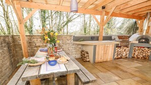 Eine Terrasse im Freien mit einem Holztisch, gedeckt für das Essen, einer Steinsäule und einem Grillbereich mit Grill und Pizzaofen.