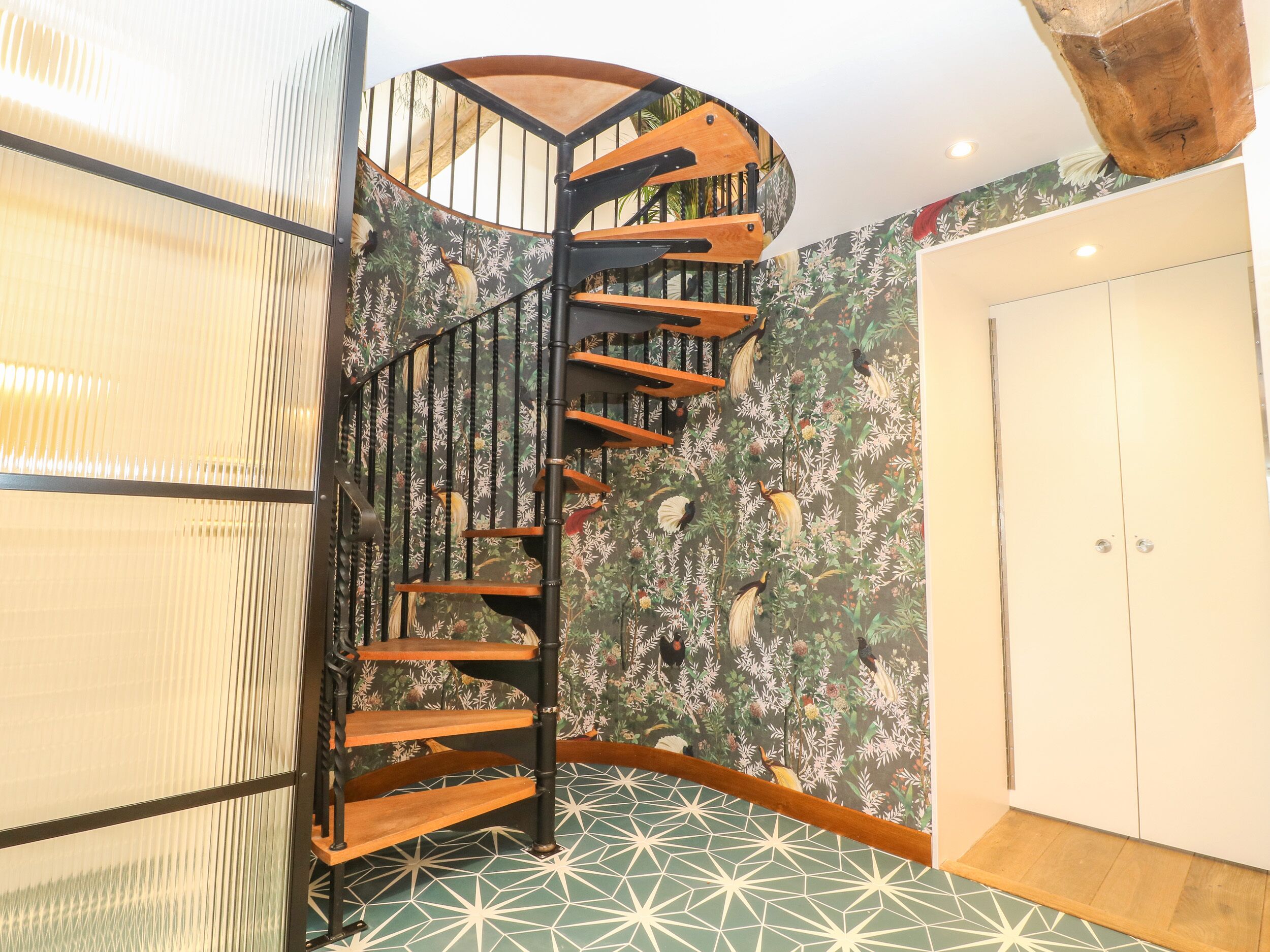A spiral staircase with wooden steps and black railings, leading to a room with a floral wallpaper and geometric patterned floor.