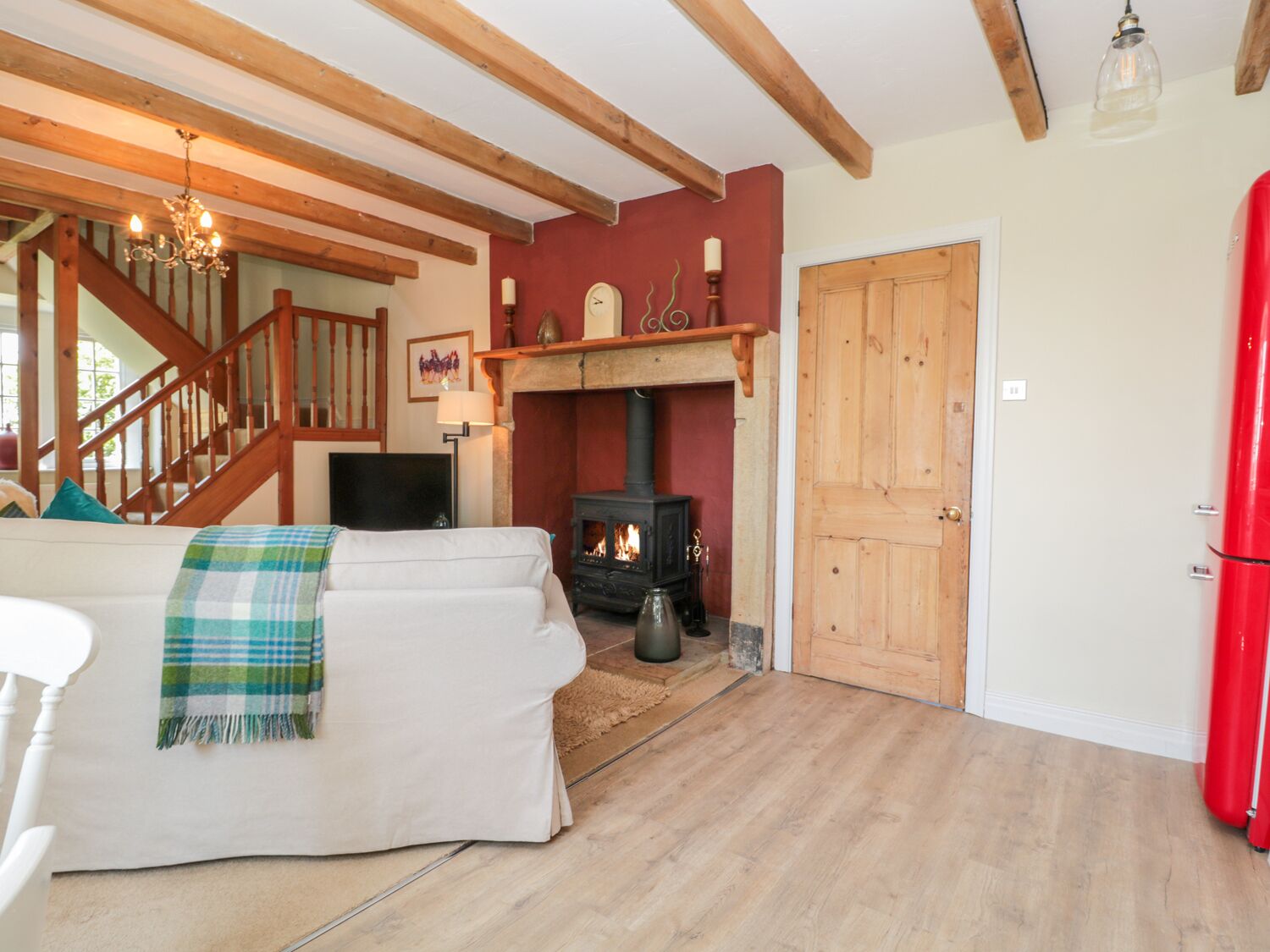 A cozy living room with a fireplace, a sofa with a plaid blanket, a wooden staircase, and a red refrigerator.