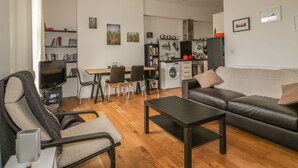 A modern living room with a sofa, a coffee table, and a TV. There is a dining area with chairs and a table. The kitchen is visible in the background with appliances and a washing machine. - Cherrywood Manor (Bodmin)