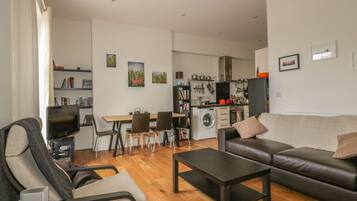 Una sala de estar moderna con un sofá, una mesa de centro y un televisor. Se observa un comedor con sillas y una mesa. En el fondo, se puede ver la cocina con electrodomésticos y una lavadora.