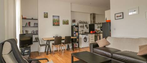 Uma sala de estar moderna com um sofá, uma mesa de centro e uma televisão. Há uma área de jantar com cadeiras e uma mesa. A cozinha é visível ao fundo, com eletrodomésticos e uma máquina de lavar.