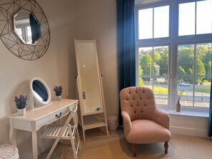 A room with a vanity, a chair, a large mirror, and a window with a view of a street and greenery. - Cherrywood Manor (Bodmin)
