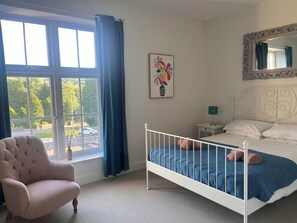 A bedroom with a bed, a chair, a nightstand, a lamp, and a window with curtains. - Cherrywood Manor (Bodmin)