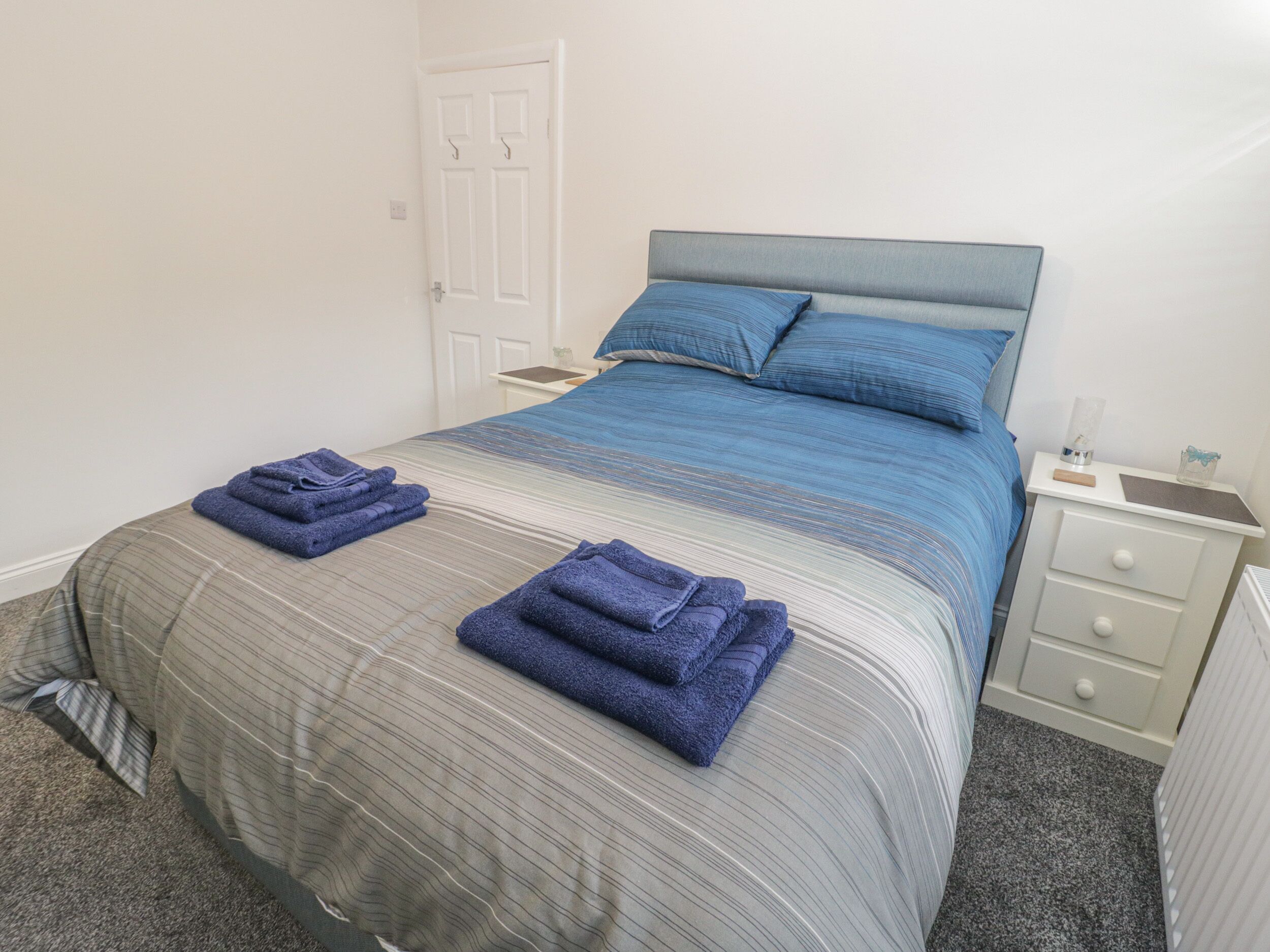 Een net opgemaakt bed met blauwe en grijze beddengoed, twee gevouwen handdoeken op het bed, een nachtkastje met een lamp, en een witte deur op de achtergrond.