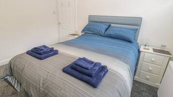Een net opgemaakt bed met blauwe en grijze beddengoed, twee gevouwen handdoeken op het bed, een nachtkastje met een lamp, en een witte deur op de achtergrond.