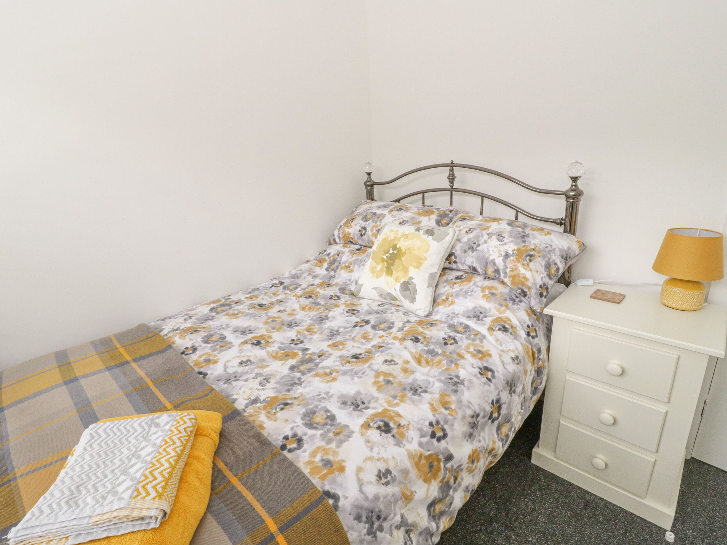 A single bed with a floral duvet, a bedside table with a lamp, and a plaid blanket.