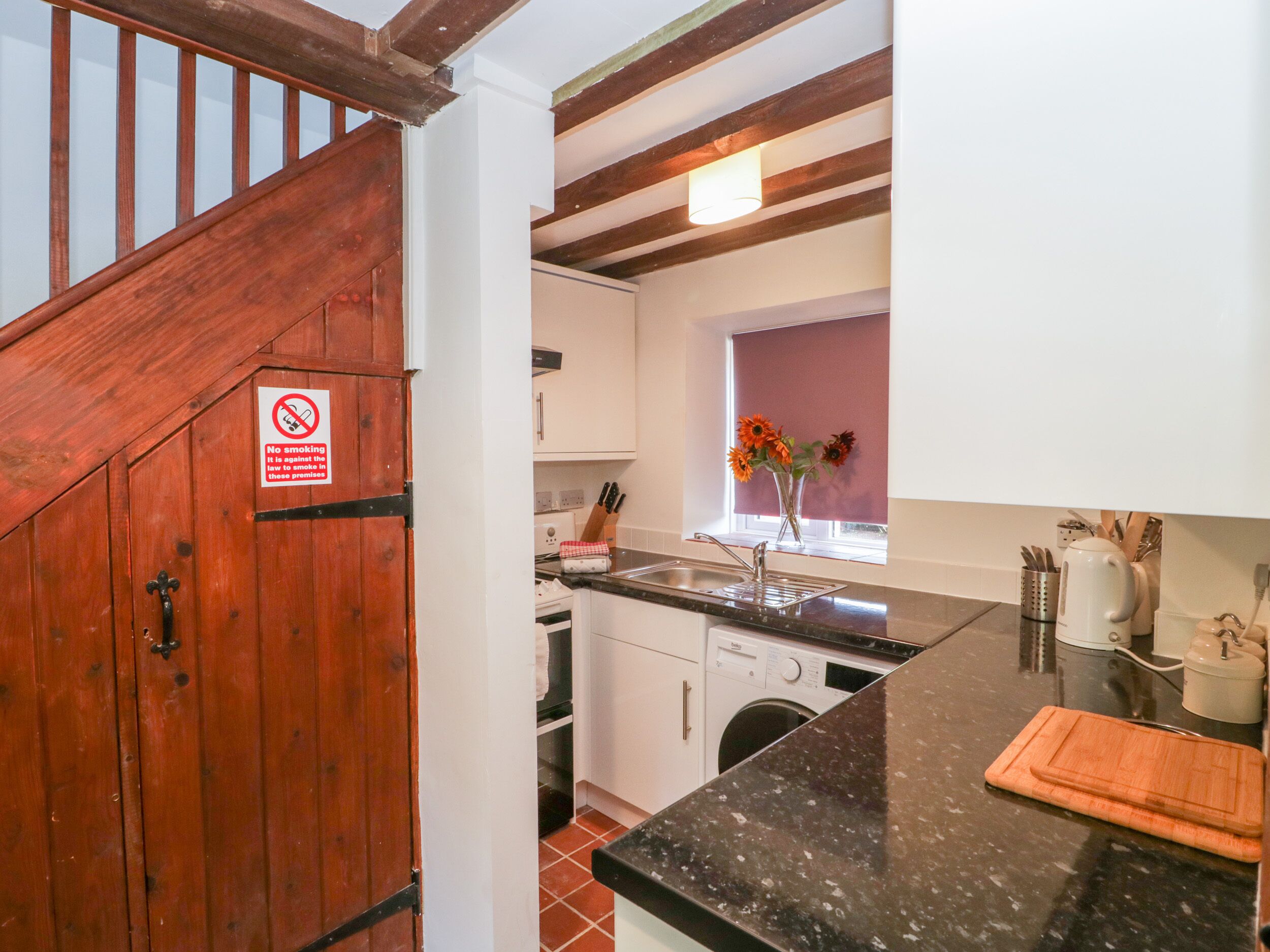 A kitchen with a wooden staircase, a washing machine, and a window with a vase of flowers.