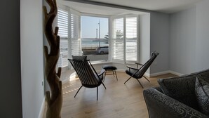 Un salon moderne doté d’une grande fenêtre donnant sur une plage, de deux fauteuils noirs et d’un canapé à motifs.