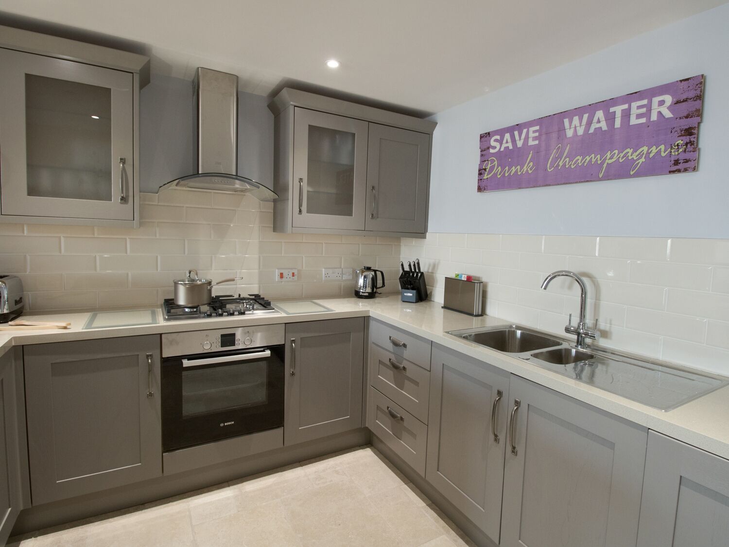 Una cocina moderna con gabinetes grises, un horno Bosch, un lavamanos y un cartel que dice 'SAVE WATER Drink champagne'.