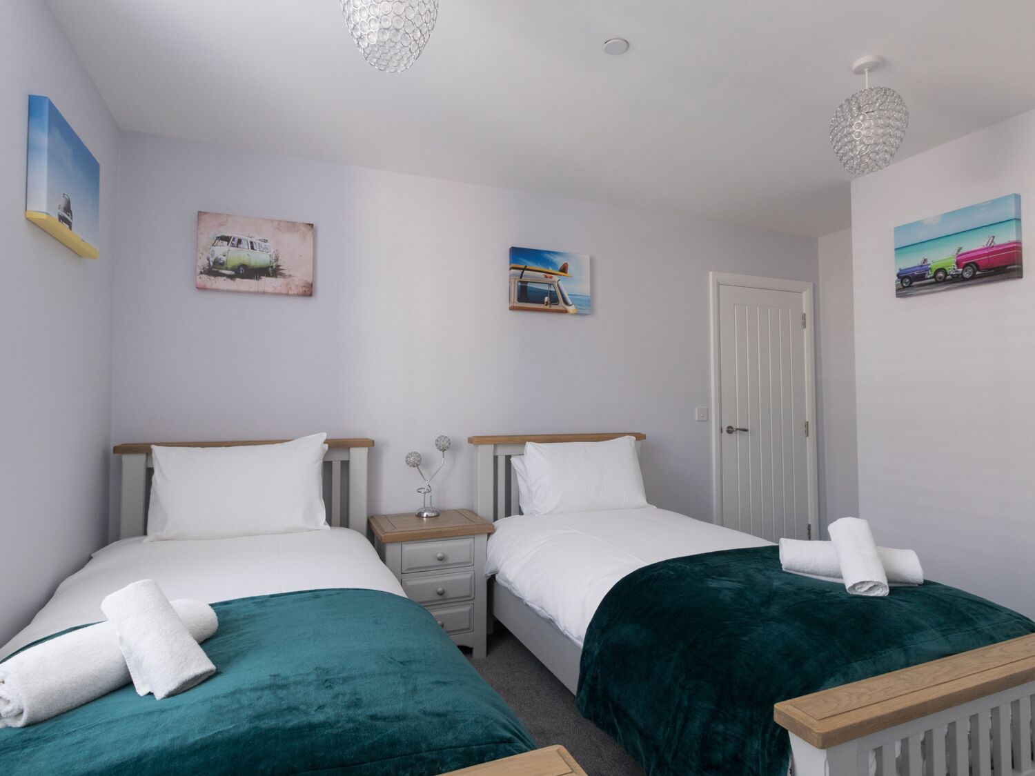 A twin room with two beds, white bedding, teal throw blankets, a bedside table with a lamp, and wall art depicting vehicles and landscapes.