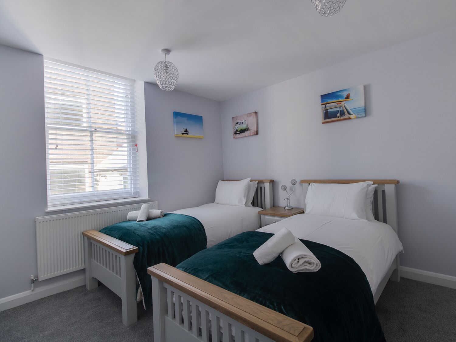 A bedroom with two single beds, a radiator, a window with blinds, and artwork on the walls.