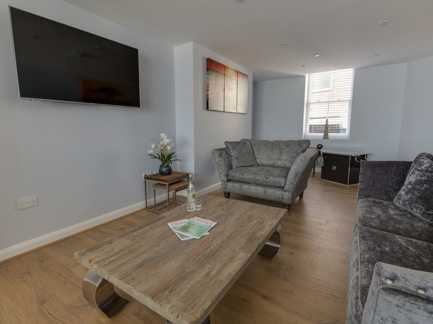 Una sala de estar con una mesa de centro de madera, un sofá gris con estampado, un televisor montado en la pared y una ventana con persianas.