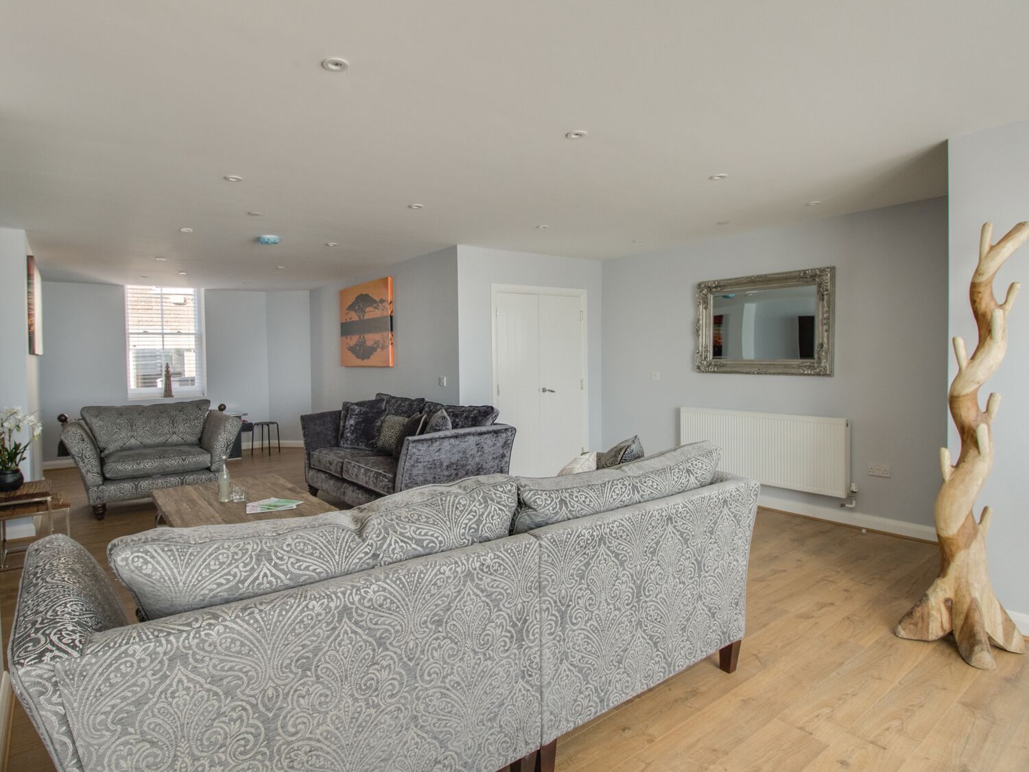 A living room with a grey patterned sofa, a smaller grey sofa, and a coffee table. There is a wooden tree sculpture on the right and a mirror on the wall.