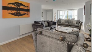 Un salon moderne avec un grand tableau, deux canapés, une table basse et une vue sur la mer grâce à de grandes fenêtres.