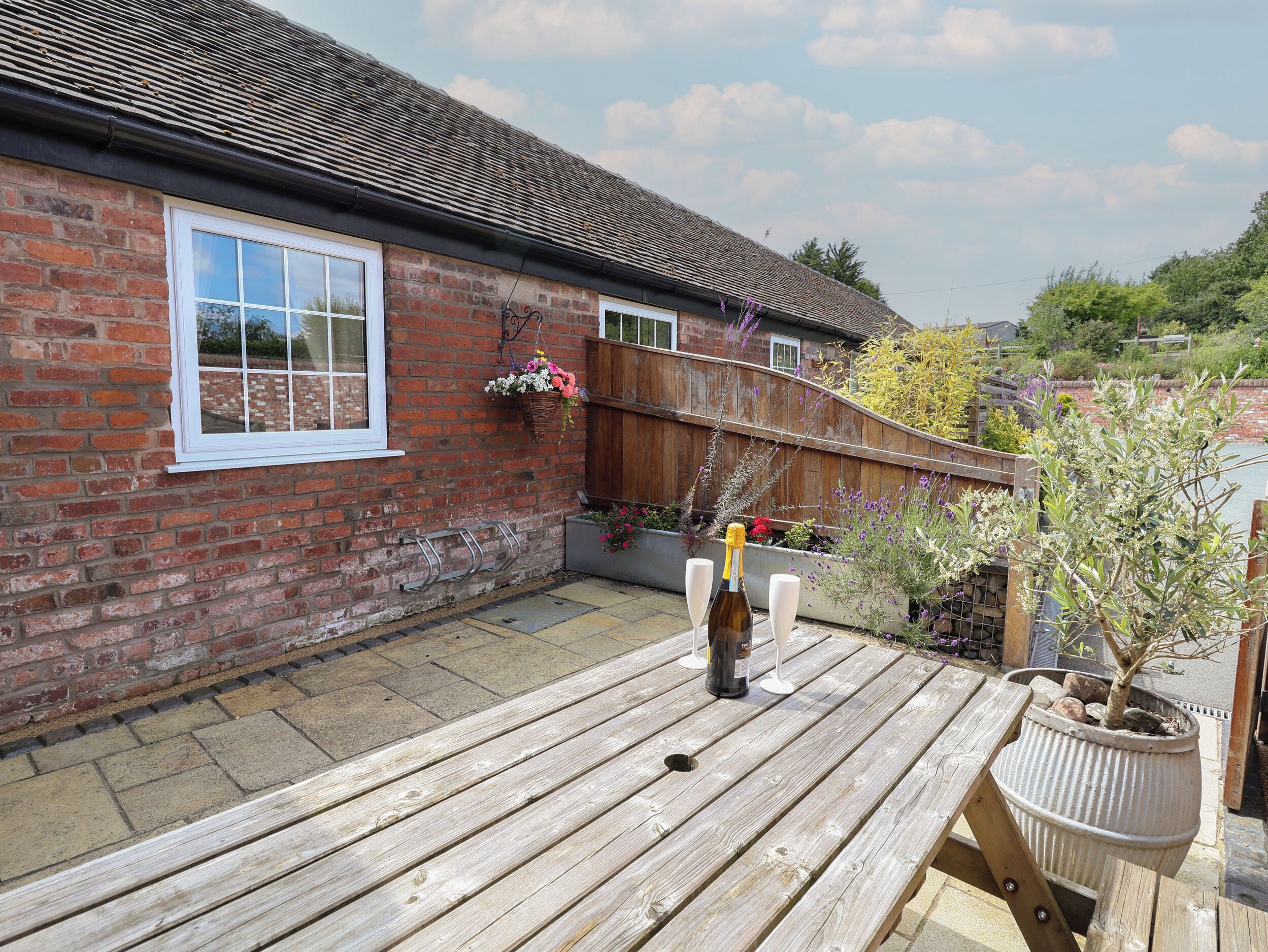 A wooden deck with a bottle of champagne and glasses, a brick house with a window, and a wooden fence.