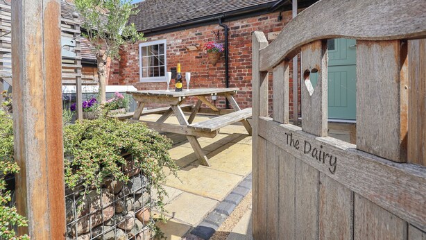 A rustic wooden gate with "The Dairy" engraved on it, leading to a brick building with a tiled roof and a window.