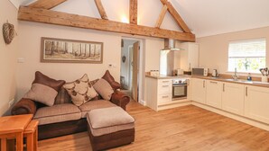A cozy living room with a wooden ceiling, a sofa with decorative pillows, a wooden coffee table, and a kitchen area with a stove, microwave, and sink.