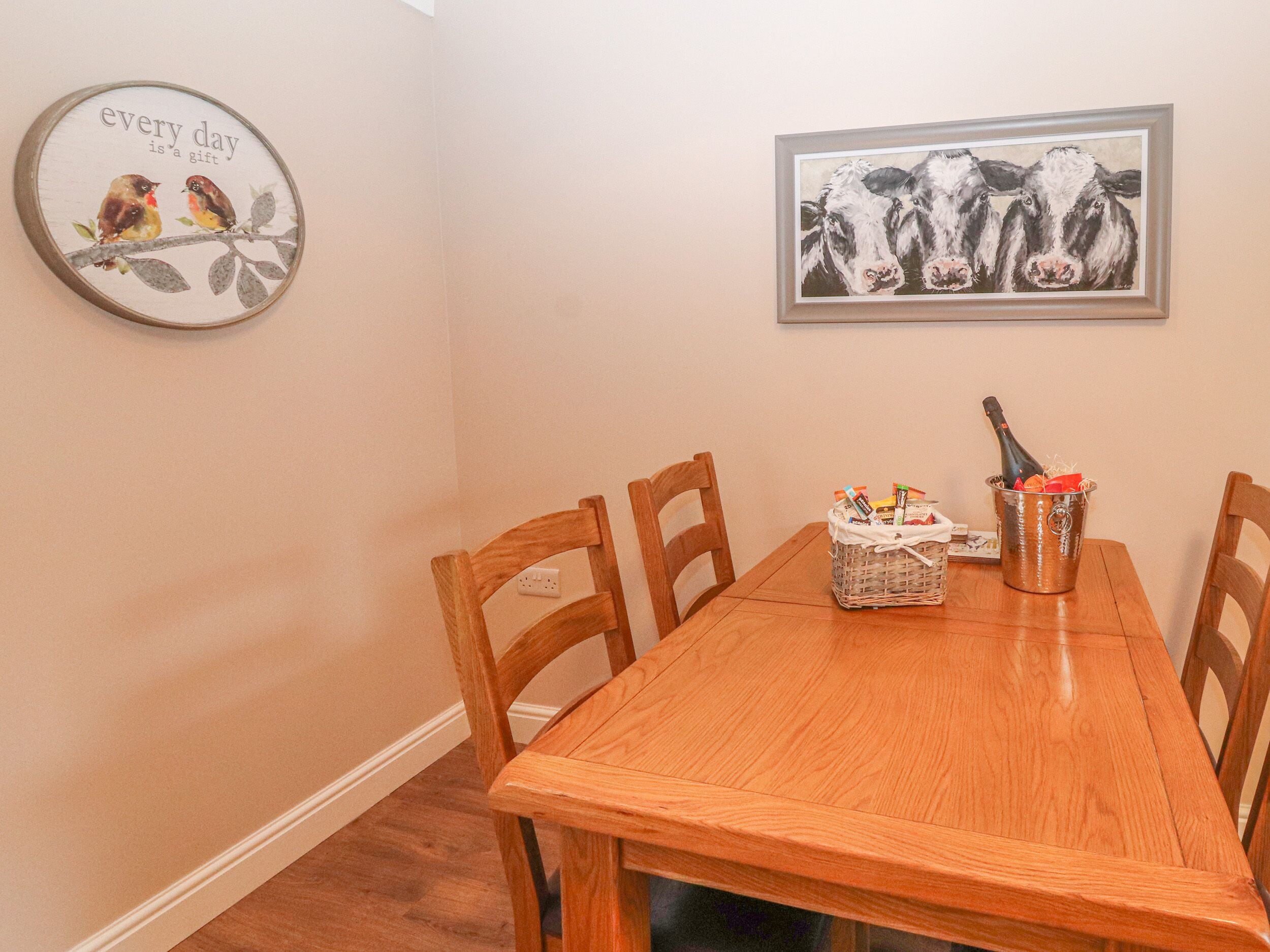Un coin repas comprenant une table en bois, quatre chaises, un tableau encadré représentant des vaches et une décoration murale avec des motifs d’oiseaux.