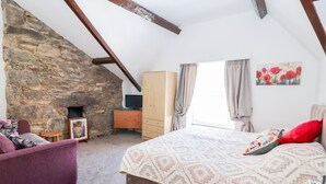 A bedroom with a stone wall, a bed with patterned bedding, a wooden dresser, a television, and a window with curtains.