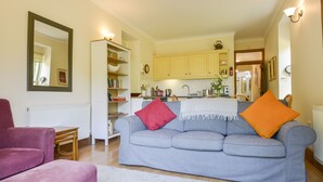 A living room with a grey sofa, a pink armchair, and a white rug. There is a kitchen in the background with yellow cabinets and a white countertop. - Engine House (Exeter)