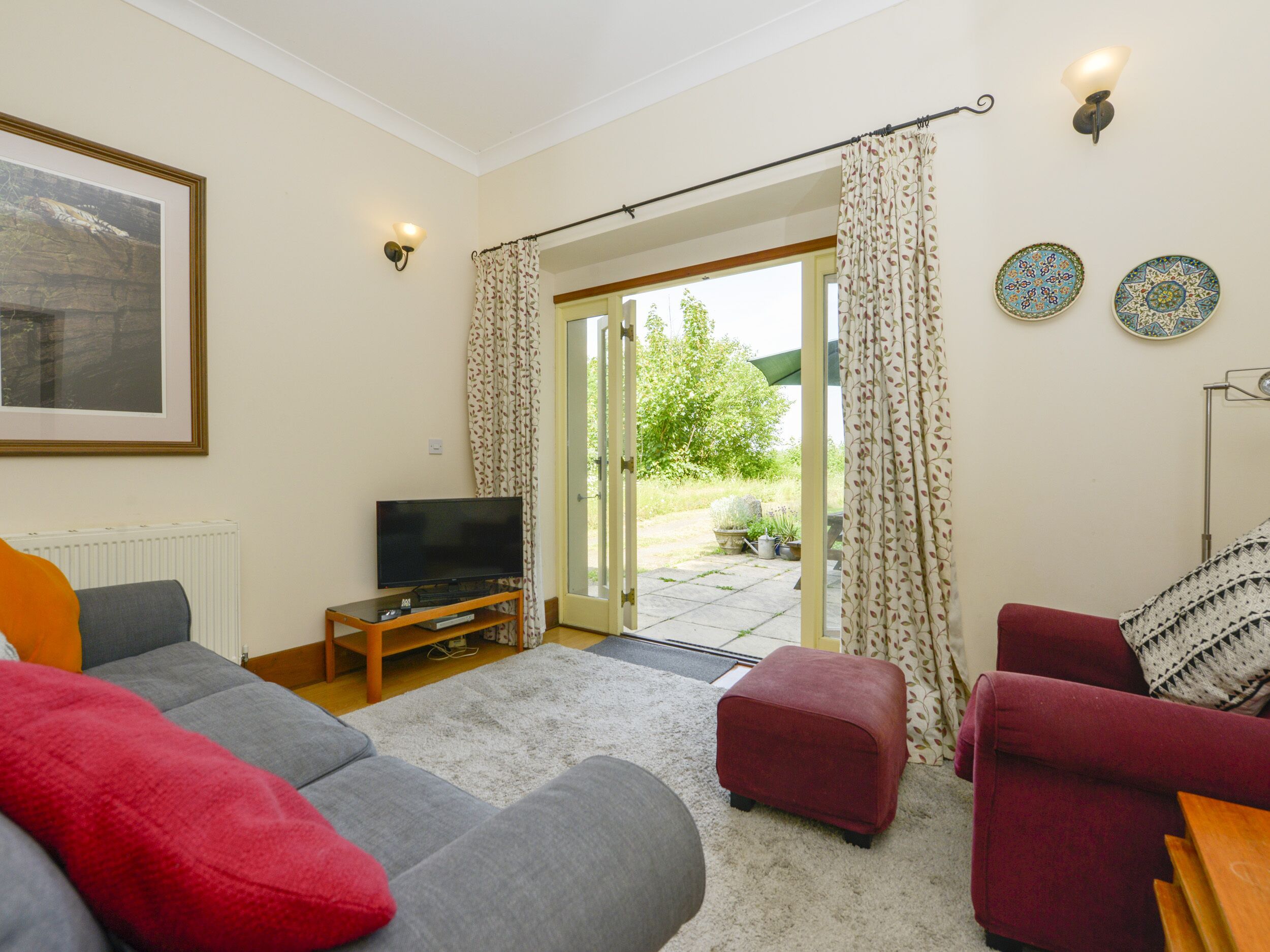 A living room with a grey sofa, a red armchair, a television, and a view of the garden through a glass door.